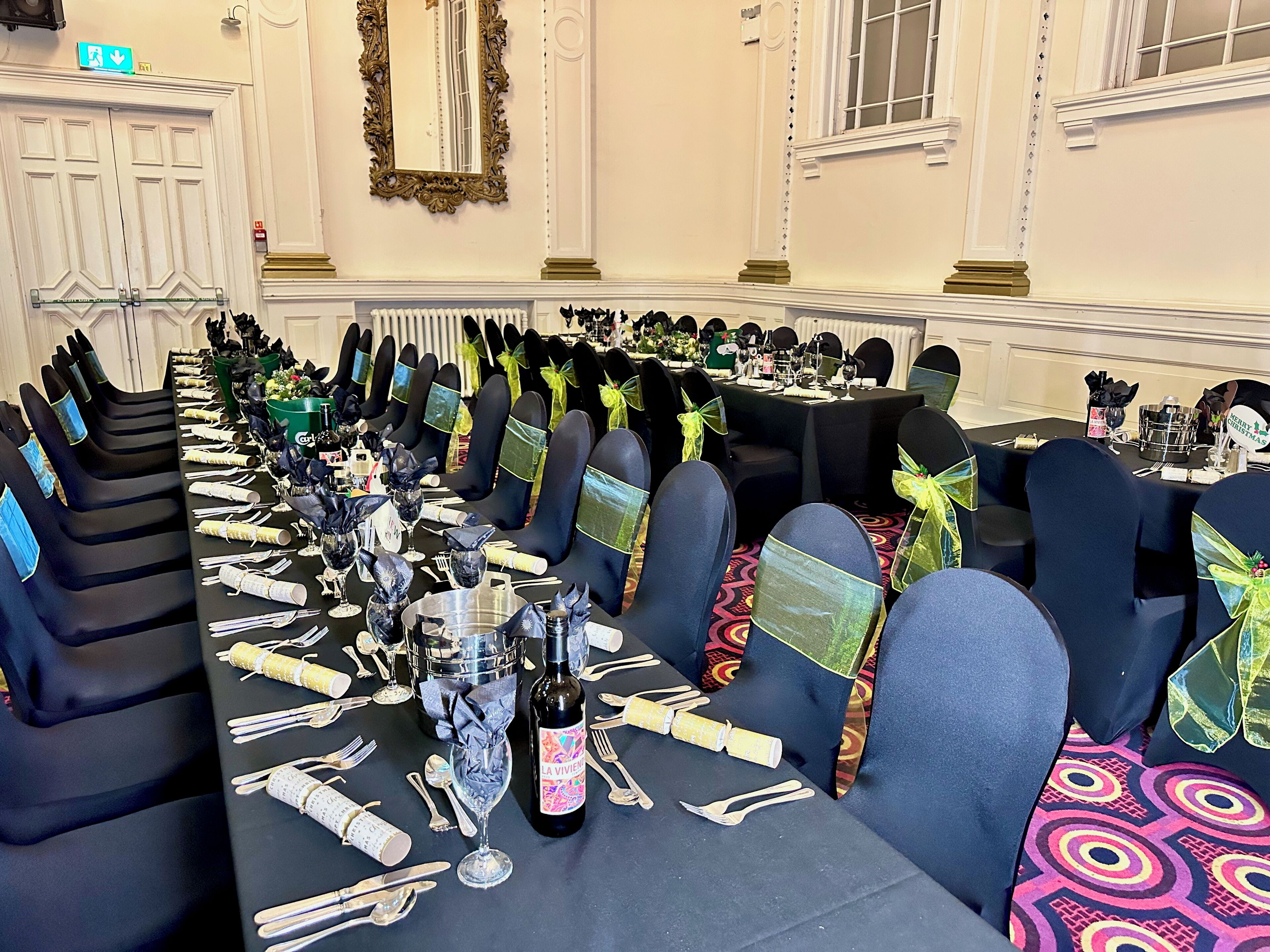 Un salón de banquetes con filas de mesas dispuestas para un evento formal, incluyendo sillas, cubiertos y una botella de vino.