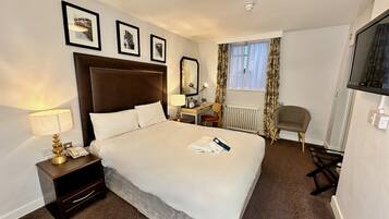A hotel room with a large bed, a desk with a chair, a TV, and framed pictures on the wall.