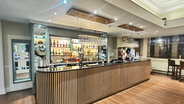 A modern bar area with a wooden counter, shelves of liquor bottles, and hanging glassware. - Carlisle Station Hotel, Sure Hotel Collection by BW (Carlisle)