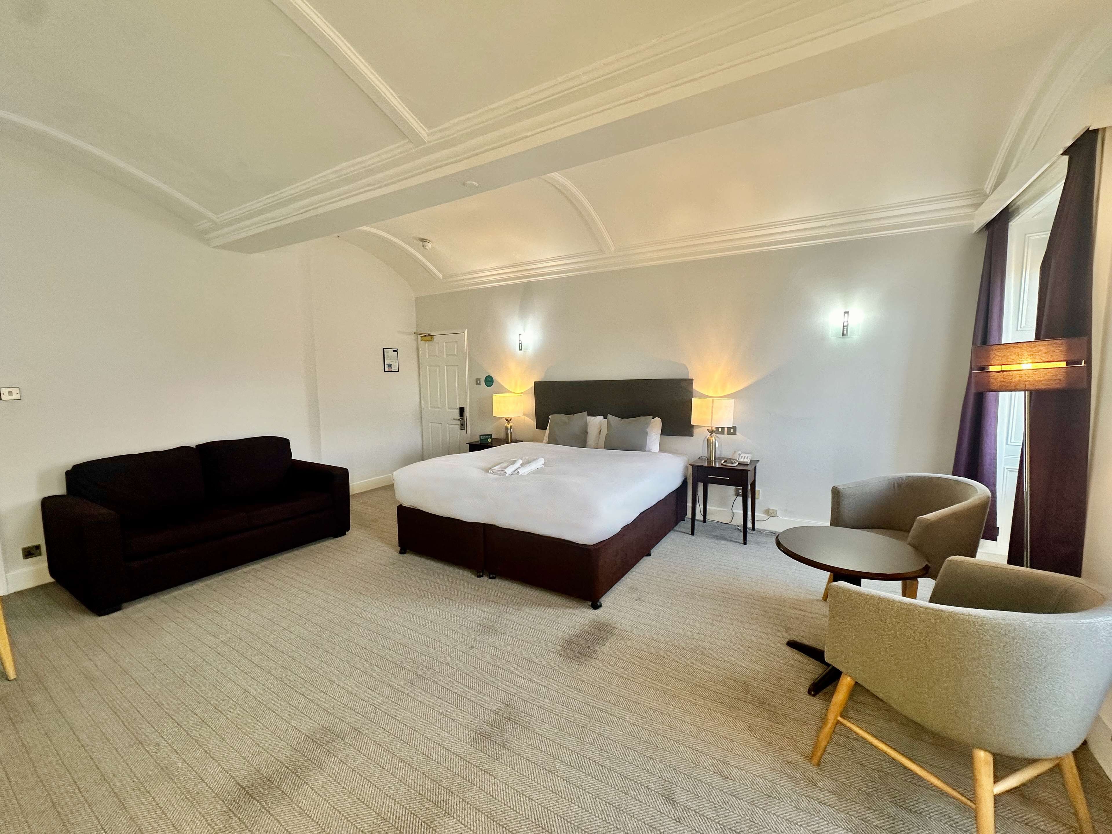 A hotel room with a large bed, a dark sofa, a small round table, and two armchairs.