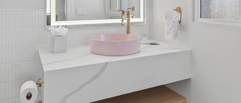 Un baño con tocador blanco, lavamanos rosa, un espejo grande con luces, toallero y ventana con vidrio esmerilado.