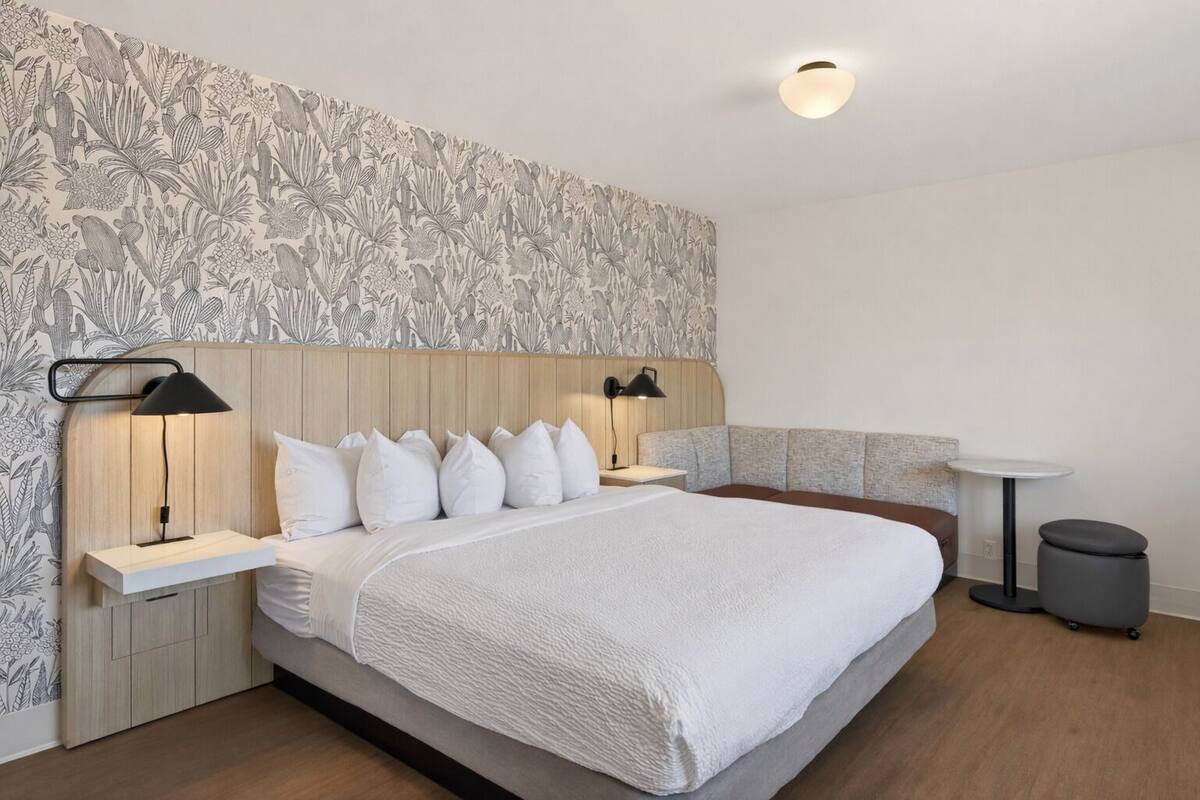 A modern hotel room with a large bed, two bedside tables with lamps, a small round table, a bench, and a trash bin.