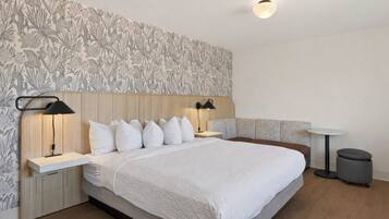 Een moderne hotelkamer met een groot bed, twee nachtkastjes met lampen, een klein rond tafeltje, een bankje en een prullenbak.