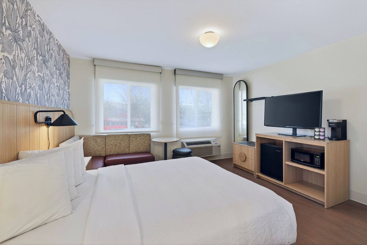 A hotel room with a large bed, a television, a minibar, and a seating area with a view of the outside.