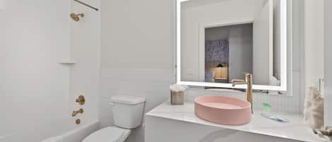 Un baño moderno con inodoro blanco, bañera, lavamanos con recipiente rosa y espejo con iluminación.