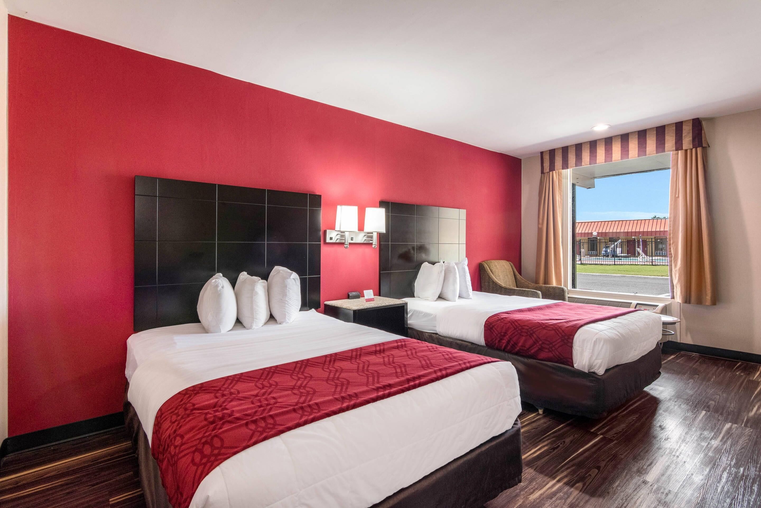 A hotel room with two beds, a large window with curtains, a headboard with a light fixture, and a view of the outside.