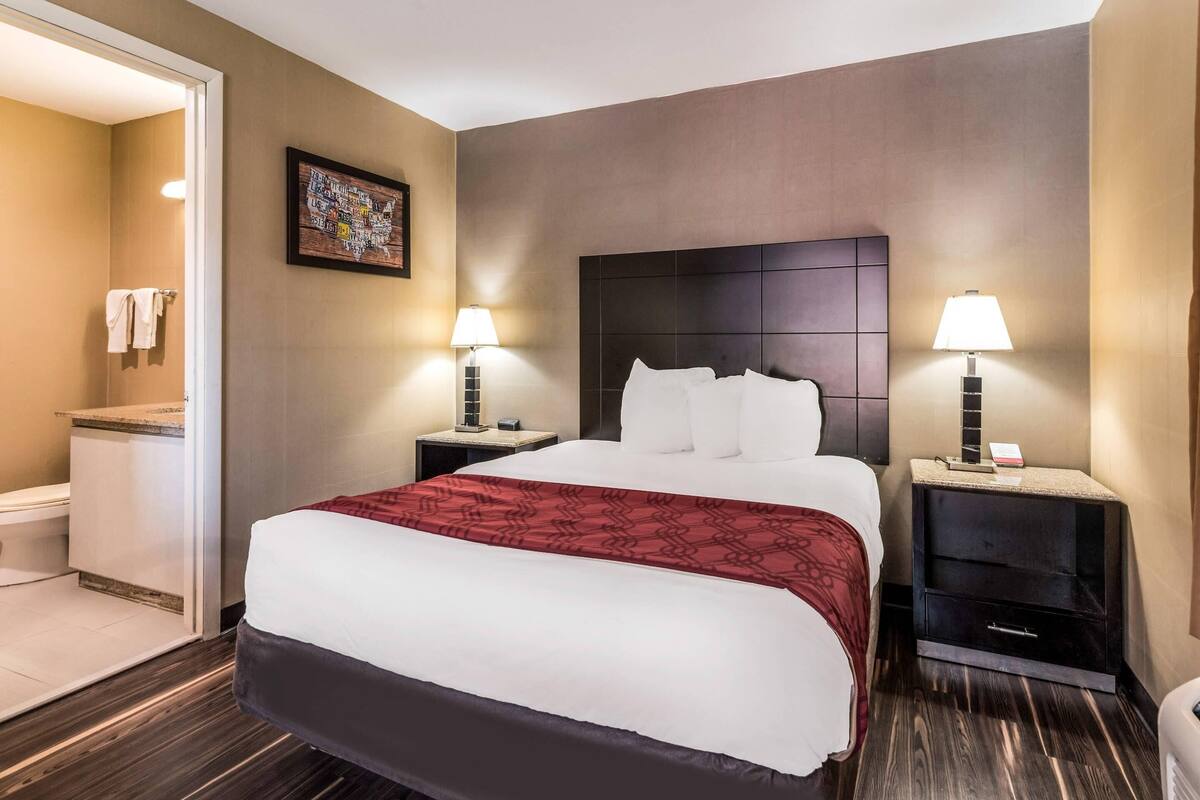 Habitación de hotel con una cama grande, dos mesitas de noche con lámparas, un cabecero, un cuadro en la pared y un baño visible a través de una puerta abierta.