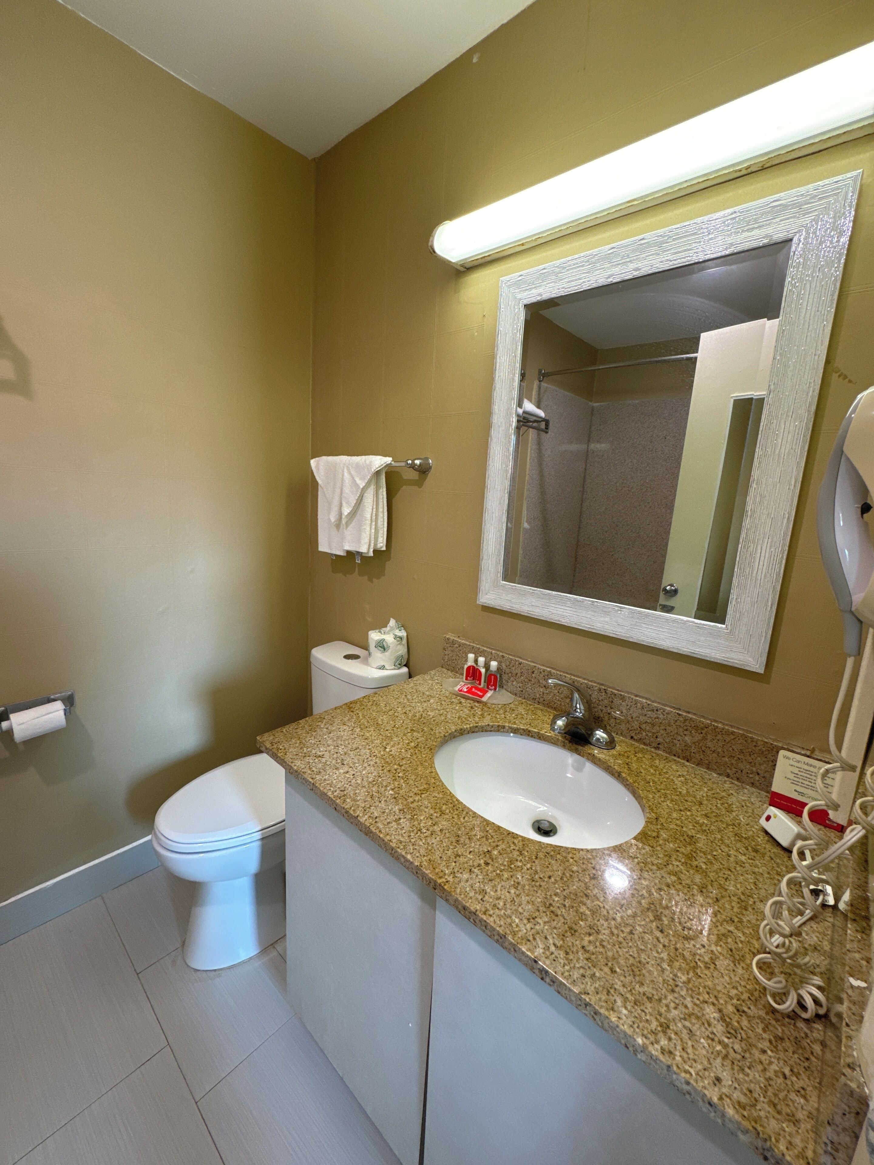 A bathroom with a white toilet, a sink with a granite countertop, a mirror, and a hand dryer.