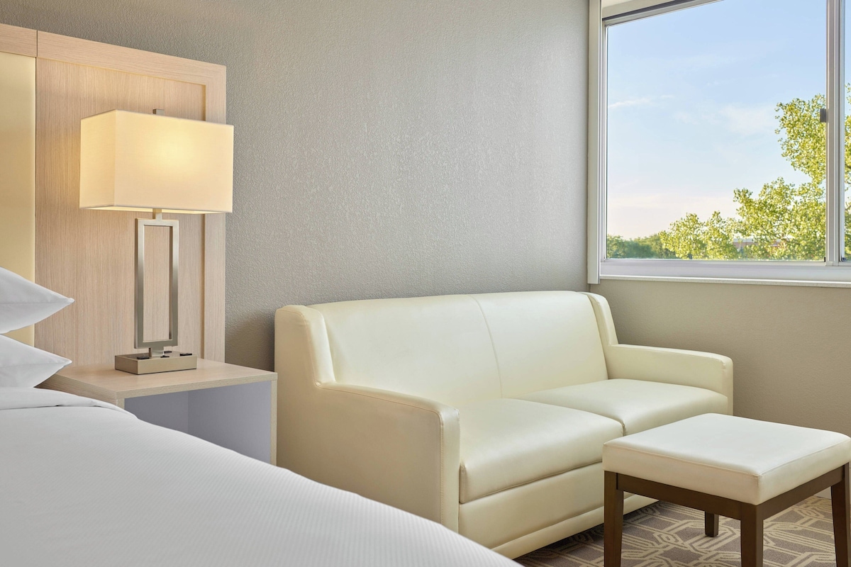A hotel room with a bed, a sofa, a small table, a lamp, and a window with a view of trees.