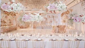 Una sala da banchetti con tavoli rotondi preparati per un evento formale, con tovaglie bianche, sedie dorate e composizioni floreali.