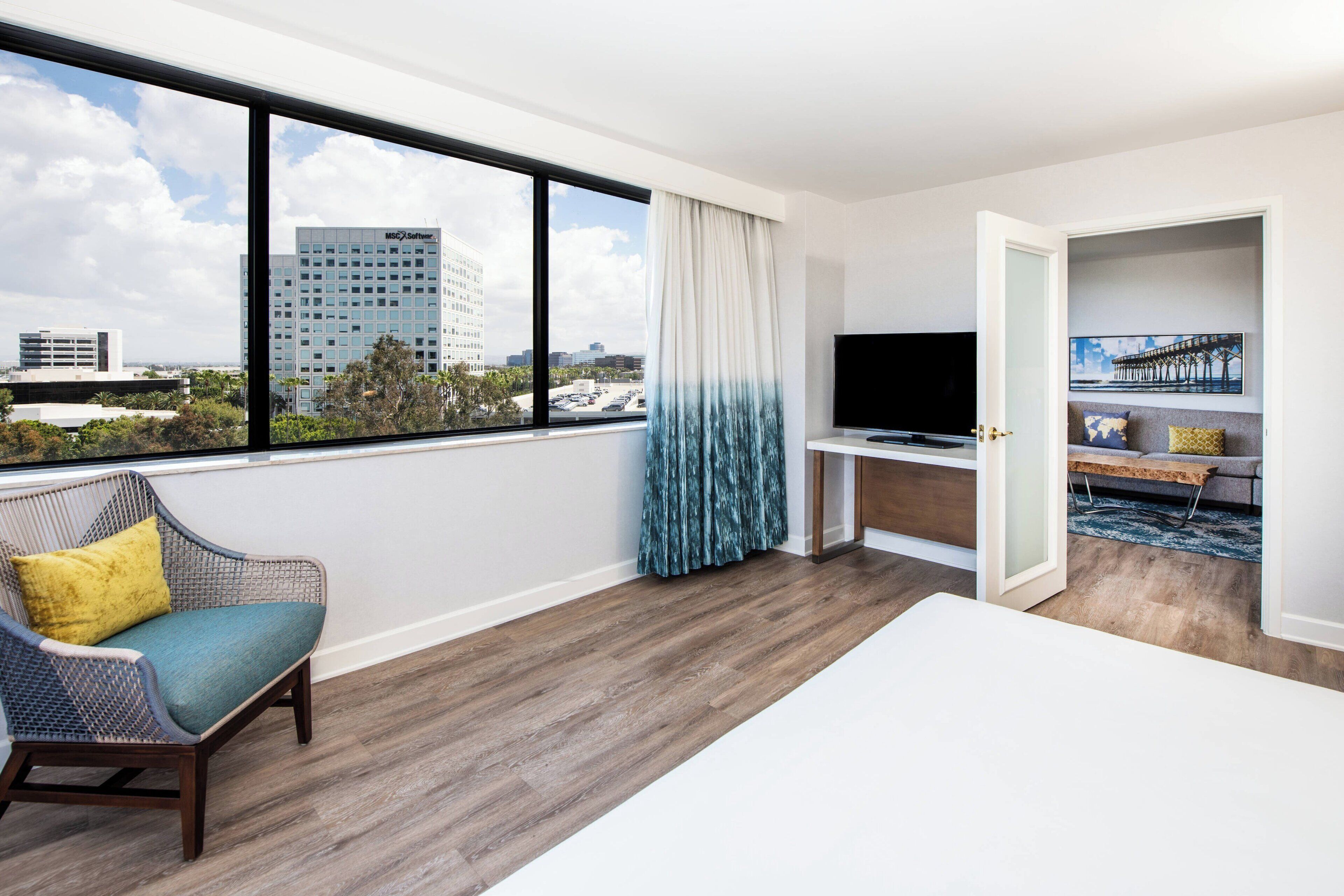 A hotel room with a large window, a flat-screen TV, a bed, a chair, and a view of buildings.