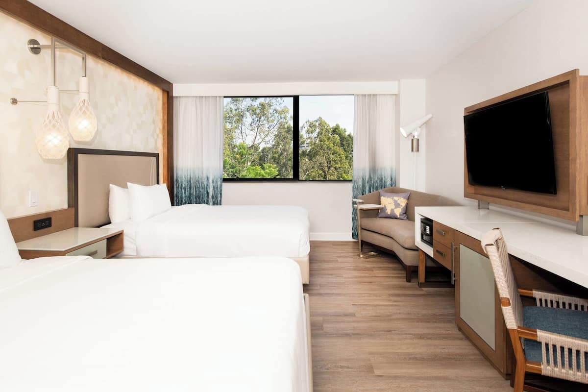 Una habitación de hotel moderna con dos camas, un televisor, un escritorio y una ventana con vista a árboles.