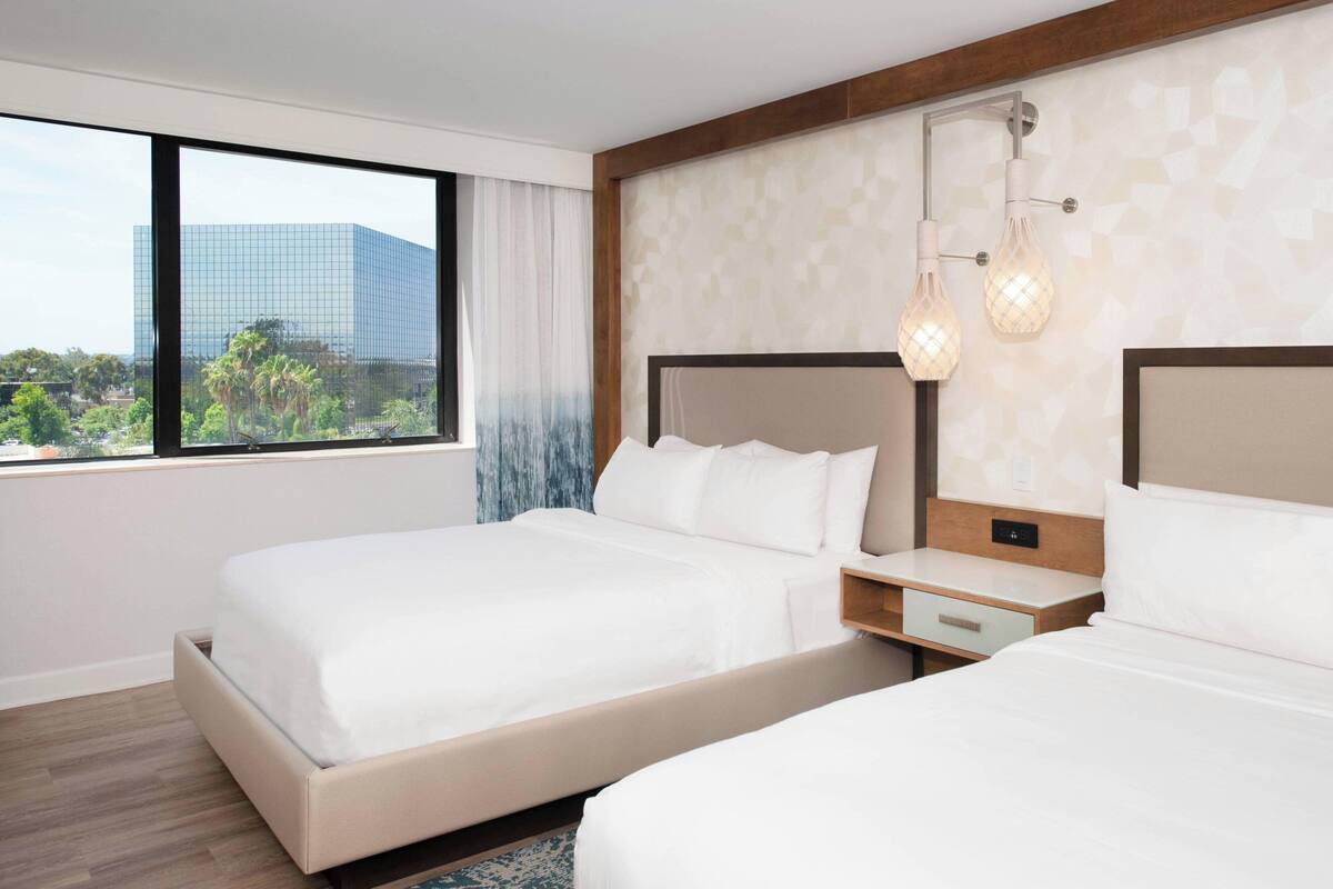 Habitación de hotel con dos camas, un ventanal amplio con vistas a la ciudad y una lámpara moderna.