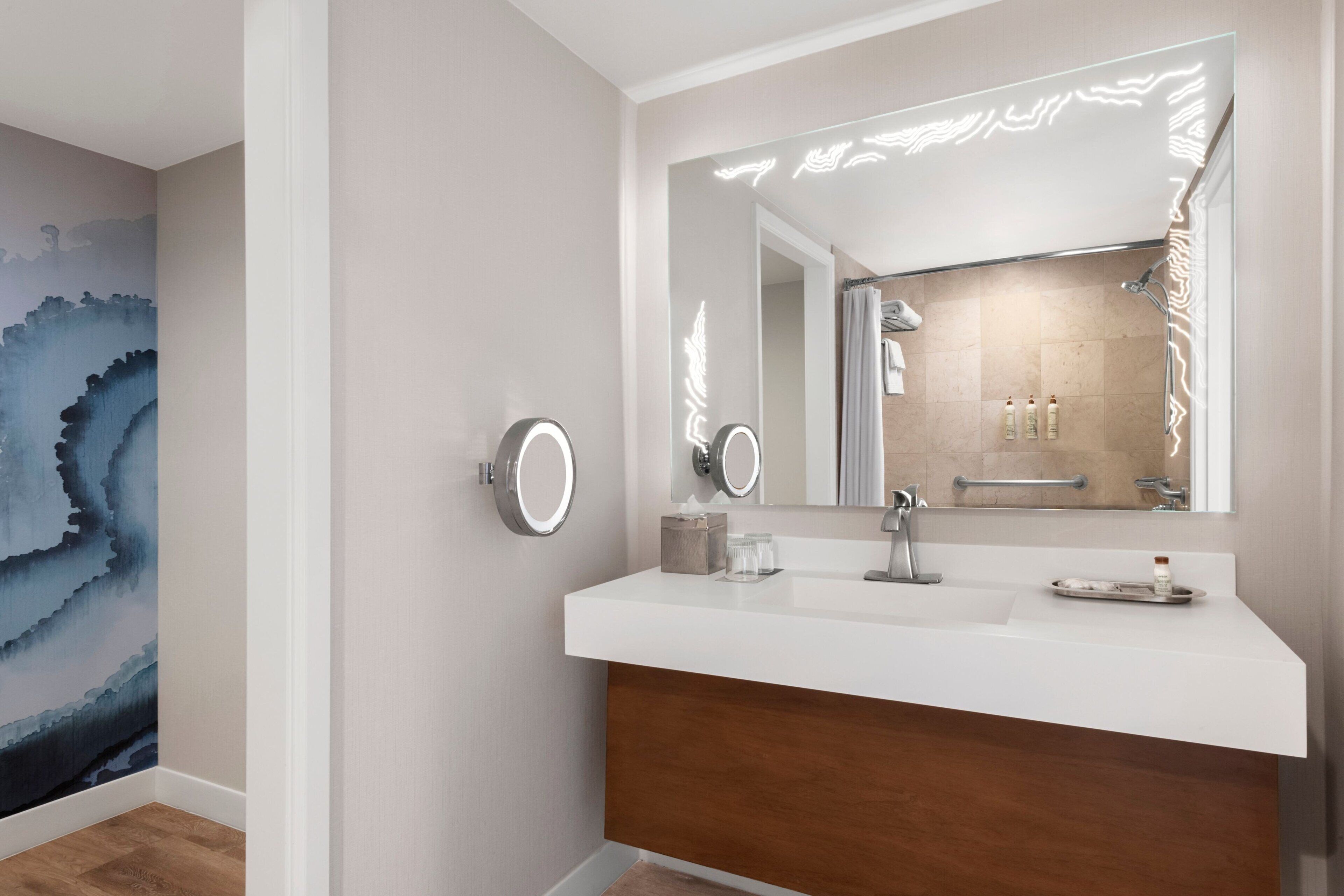 Un bany modern amb un lavabo blanc, un mirall gran amb il·luminació integrada i un armari de fusta a la part inferior.