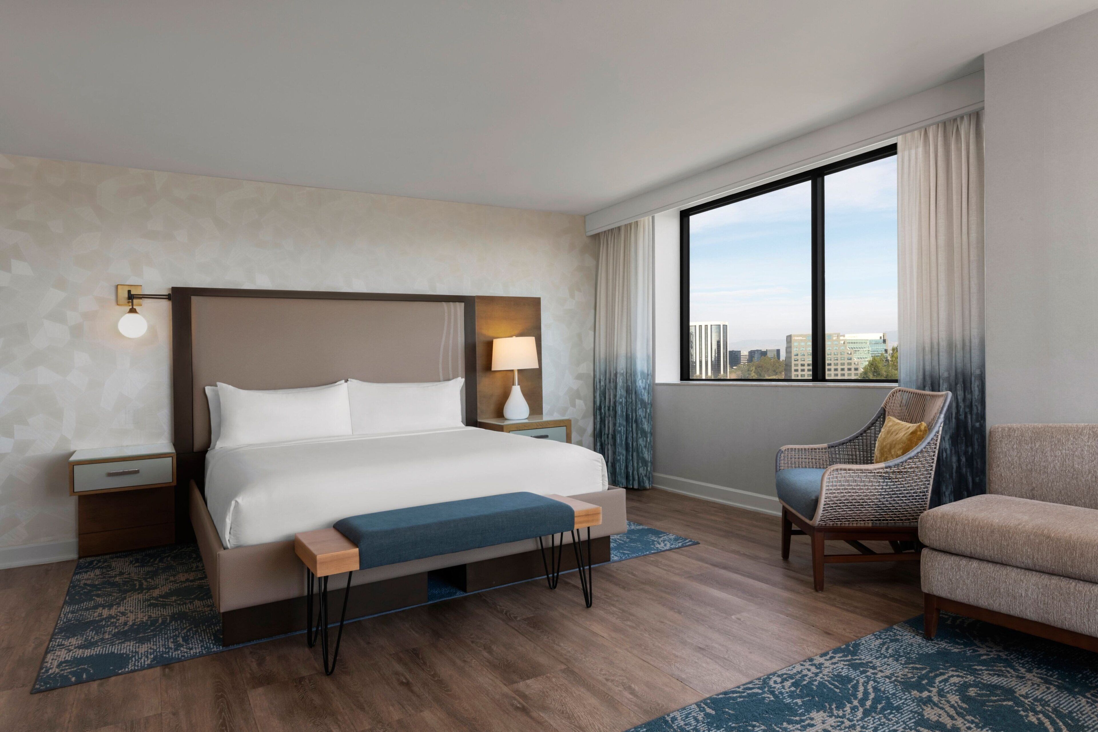 A modern hotel room with a large bed, a nightstand, a chair, and a view of the city through large windows.