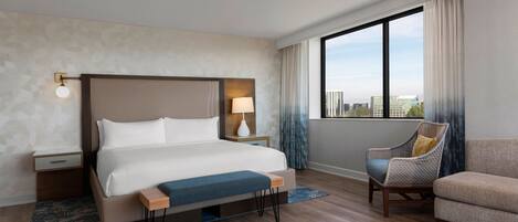 A modern hotel room with a large bed, a nightstand, a chair, and a view of the city through large windows.