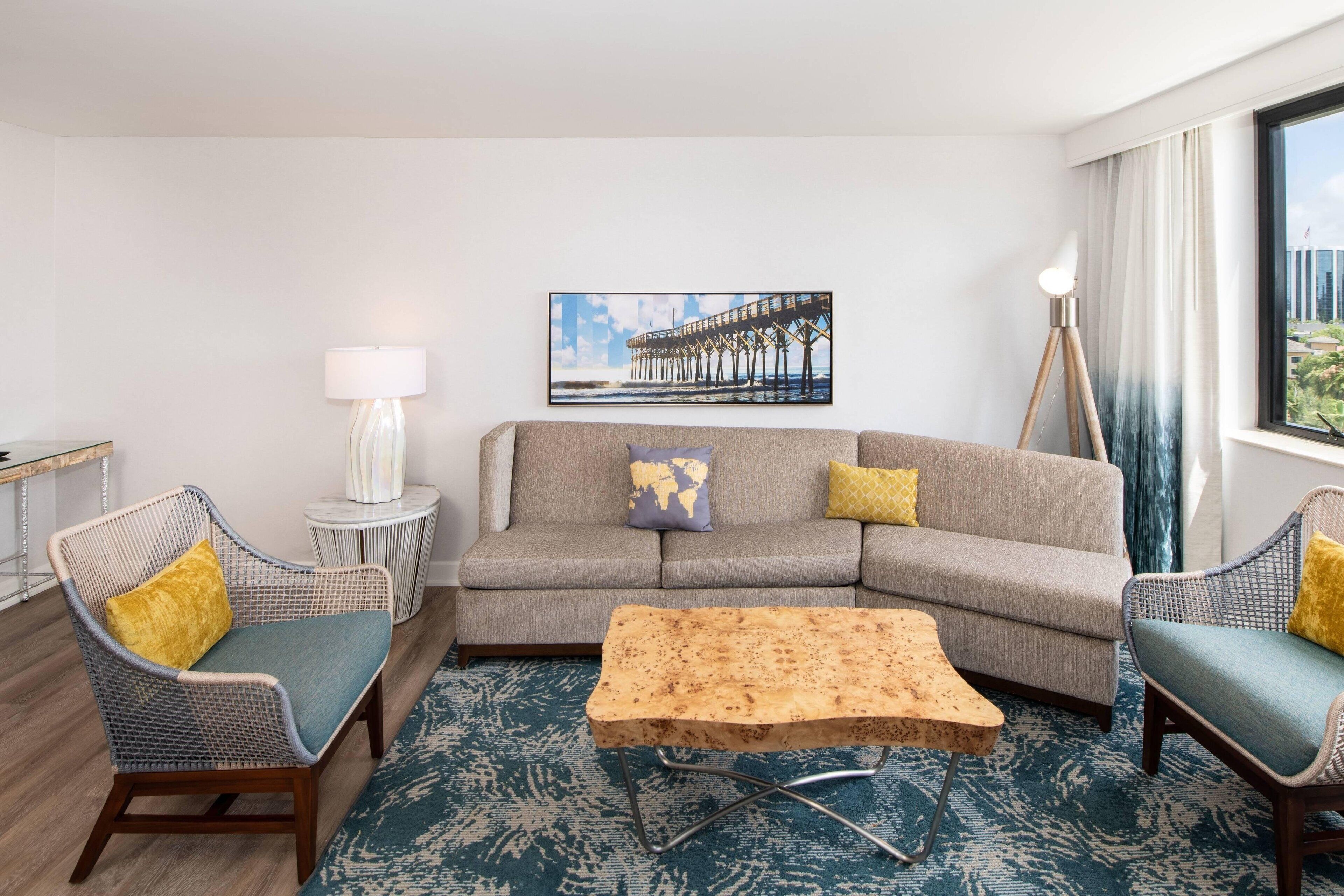 Een woonkamer met een bank, twee fauteuils, een salontafel, een lamp, een sierkussen met een wereldkaart en een foto van een pier.
