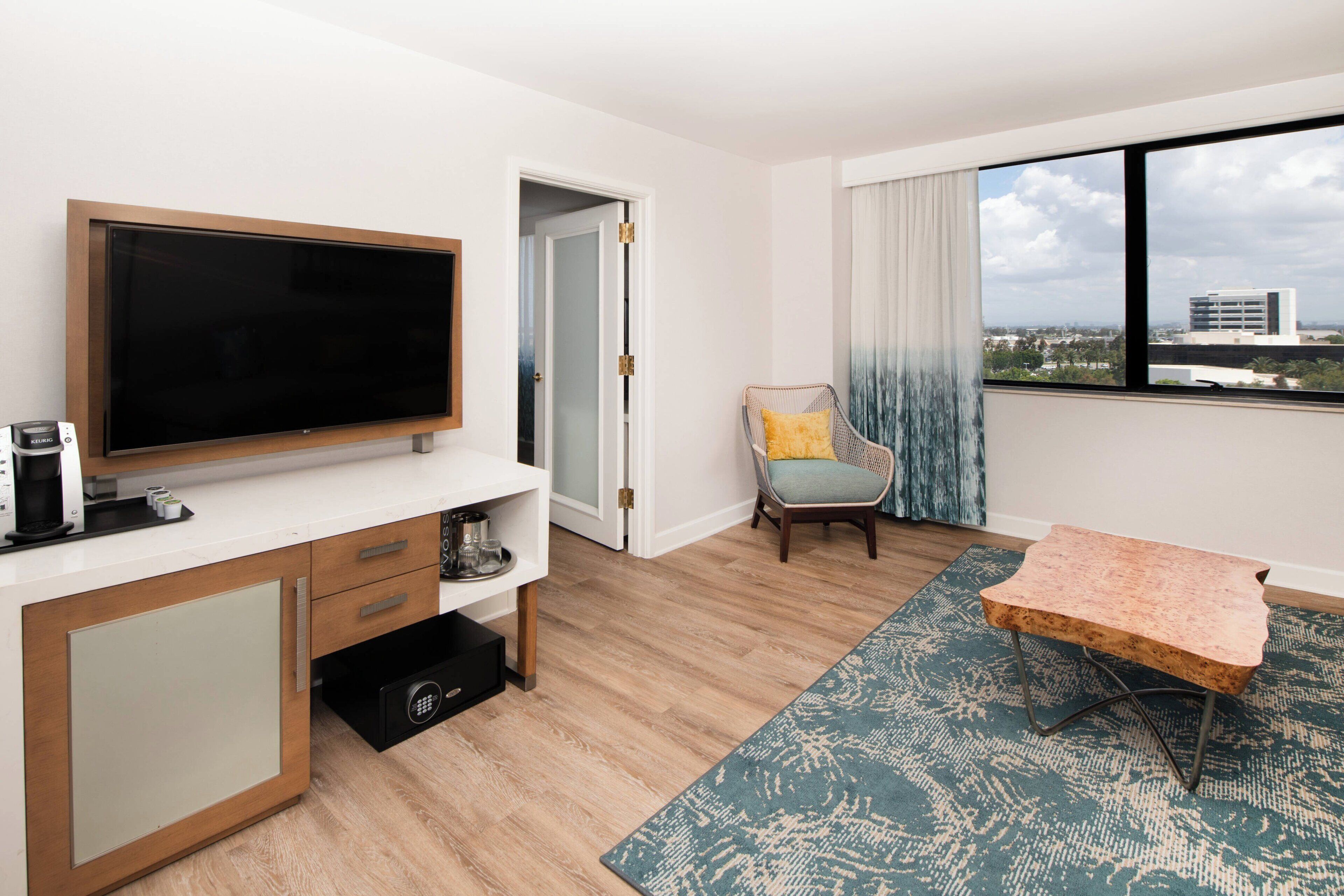 A modern hotel room with a flat-screen TV, a wooden cabinet, a small table, a chair with a cushion, and a view of the city through a large window.