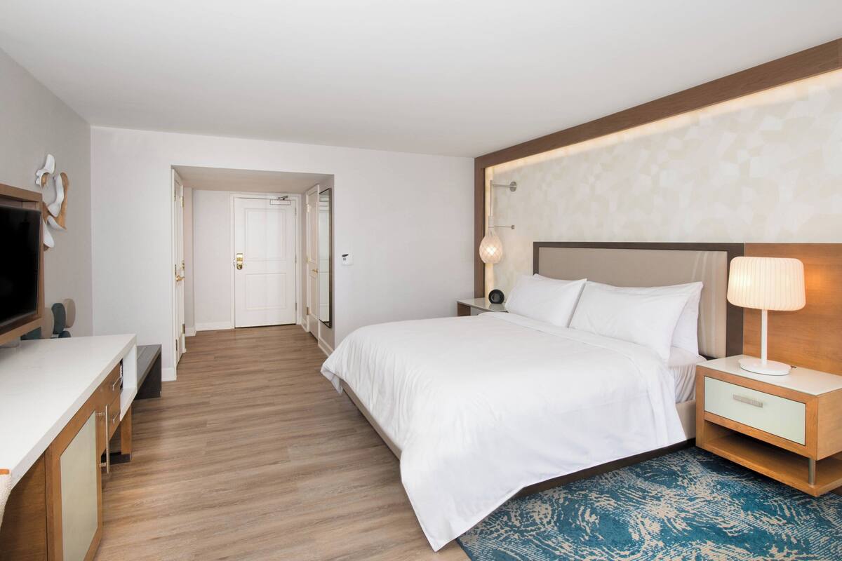 Habitación de hotel con cama, mesita de noche, lámpara y televisor sobre un escritorio.