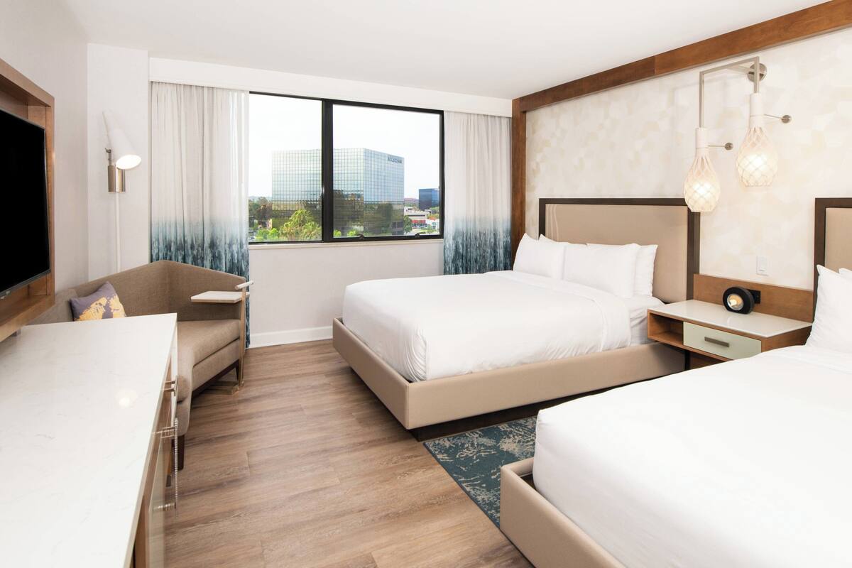 Habitación de hotel con dos camas, un escritorio, una silla, un televisor y un ventanal con vista a la ciudad.