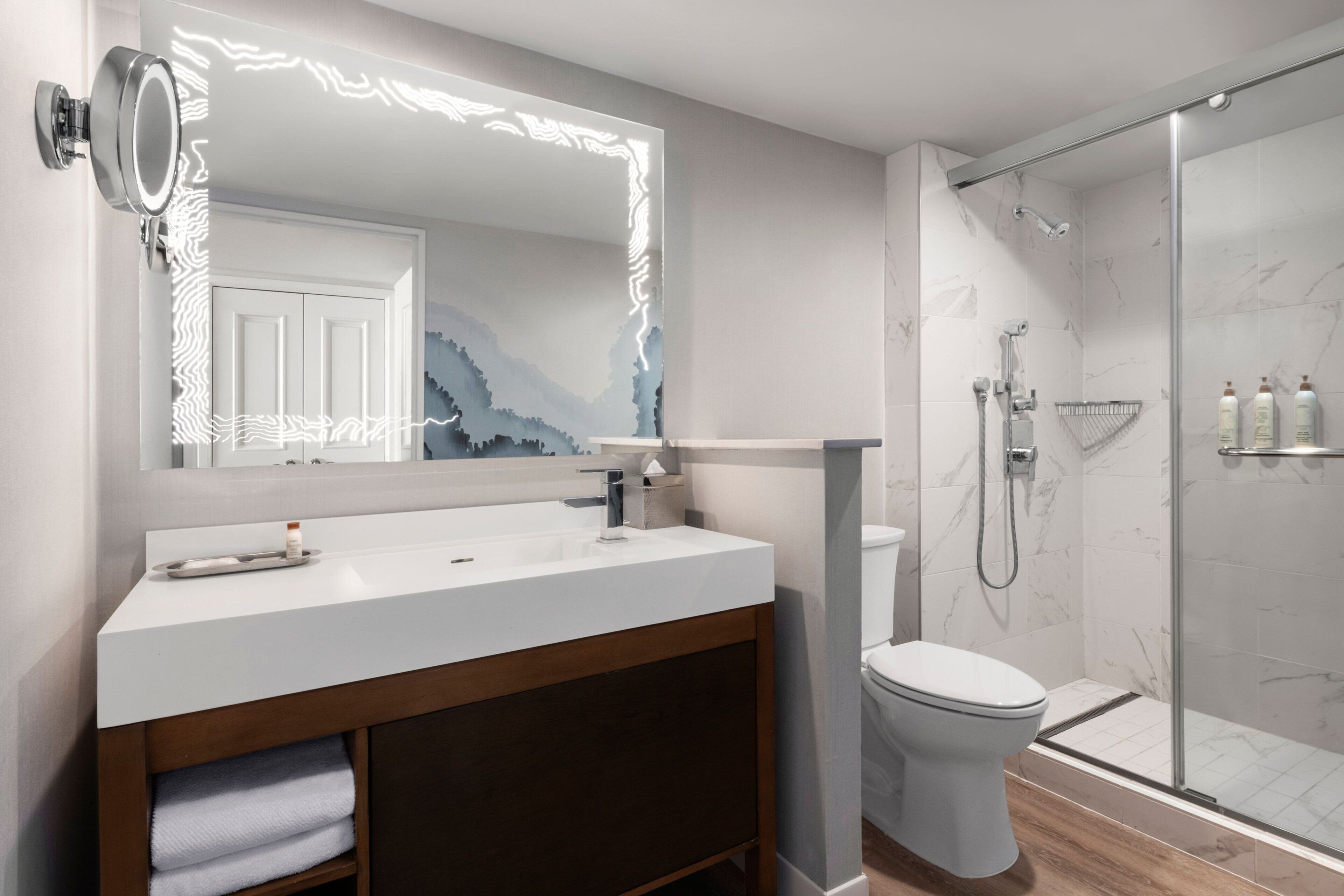 A modern bathroom with a large mirror, a white sink, a toilet, and a glass-enclosed shower.