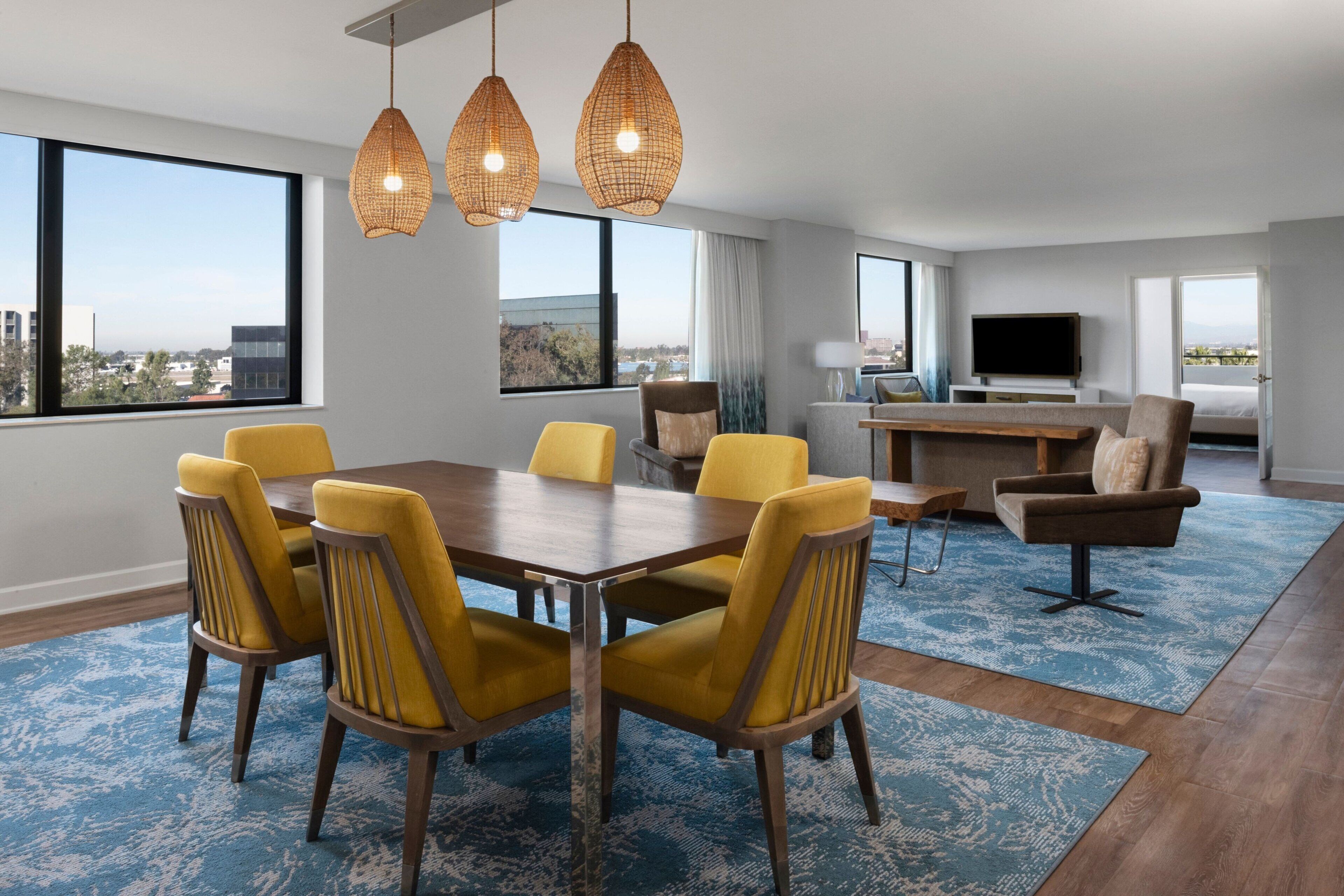 Una habitación de hotel moderna con zona de comedor, televisión y ventanas amplias con vistas a la ciudad.