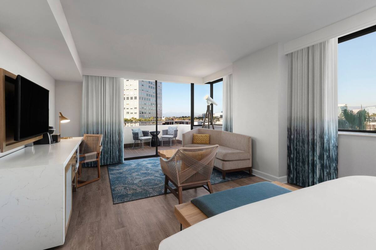 Habitación de hotel moderna con una cama grande, un sofá, una mesa de comedor y un balcón con vista a la ciudad.