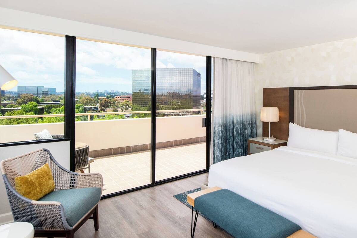 Habitación de hotel con una cama grande, un balcón con vista a la ciudad, una silla de mimbre y una pequeña mesa.