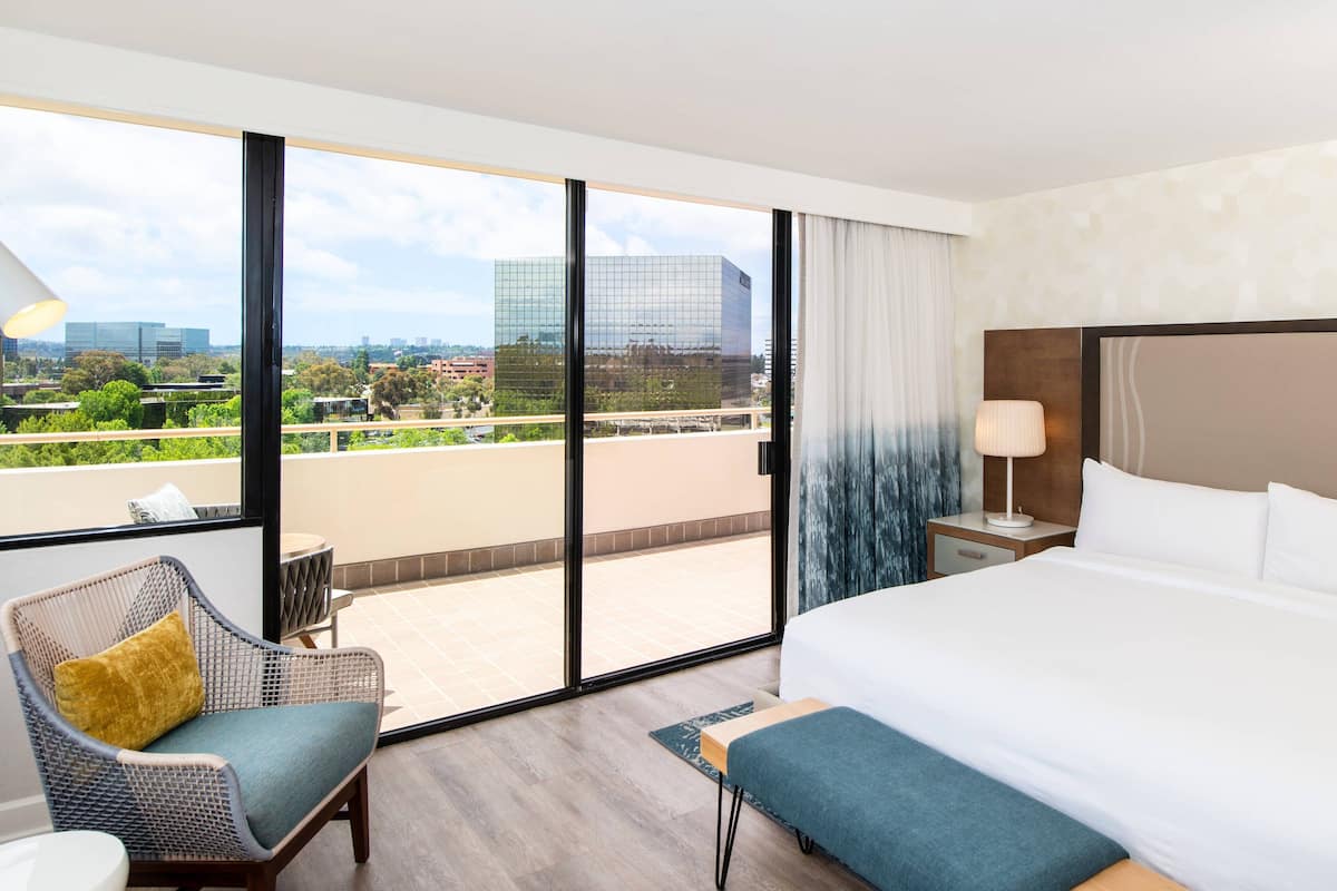 A hotel room with a large bed, a balcony with a view of the city, a wicker chair, and a small table.