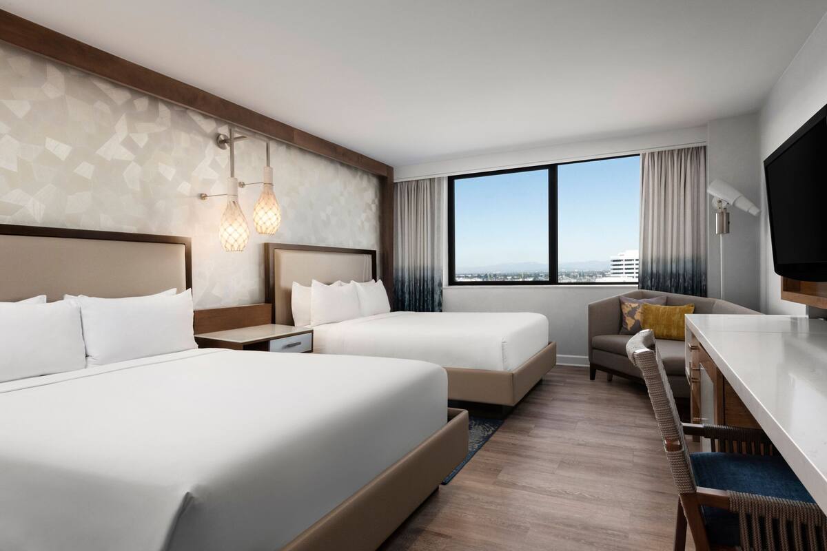 Habitación de hotel con dos camas, un escritorio, una silla, televisión y un ventanal con vista a la ciudad.