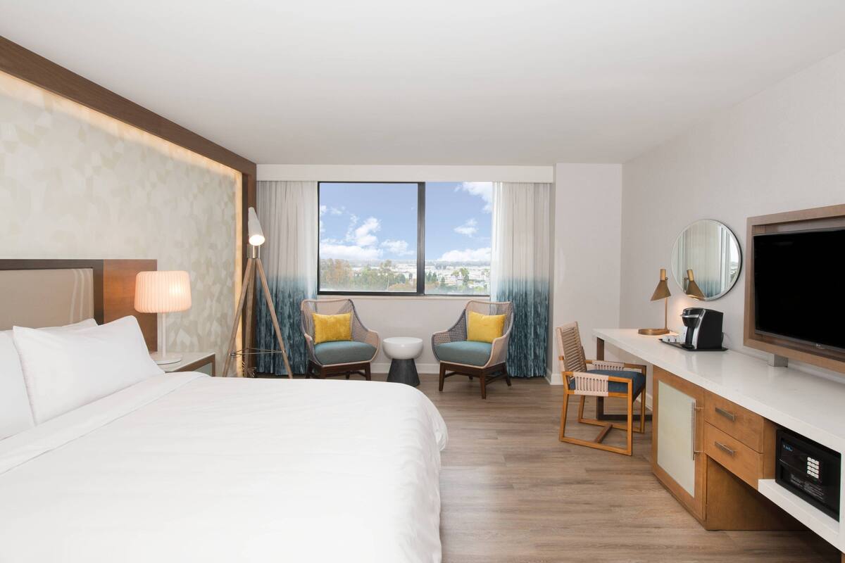 Habitación de hotel con una cama grande, un escritorio con televisor, una mesita, dos sillas y vistas a la ciudad a través de la ventana.