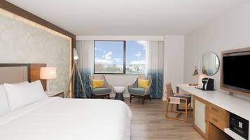 Quarto de hotel com cama, duas cadeiras, escrivaninha e televisão, além de uma janela com vista.