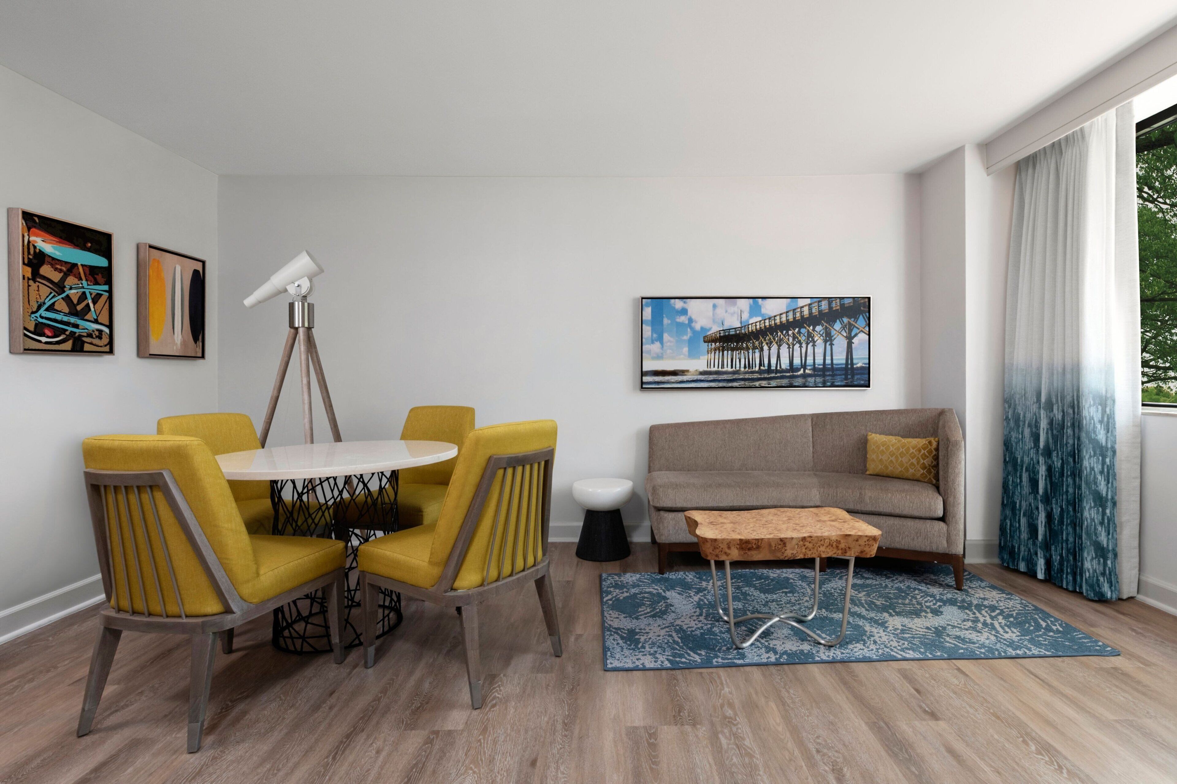 A modern living room with a dining area, featuring a round table, yellow chairs, a sofa, and a painting of a pier.