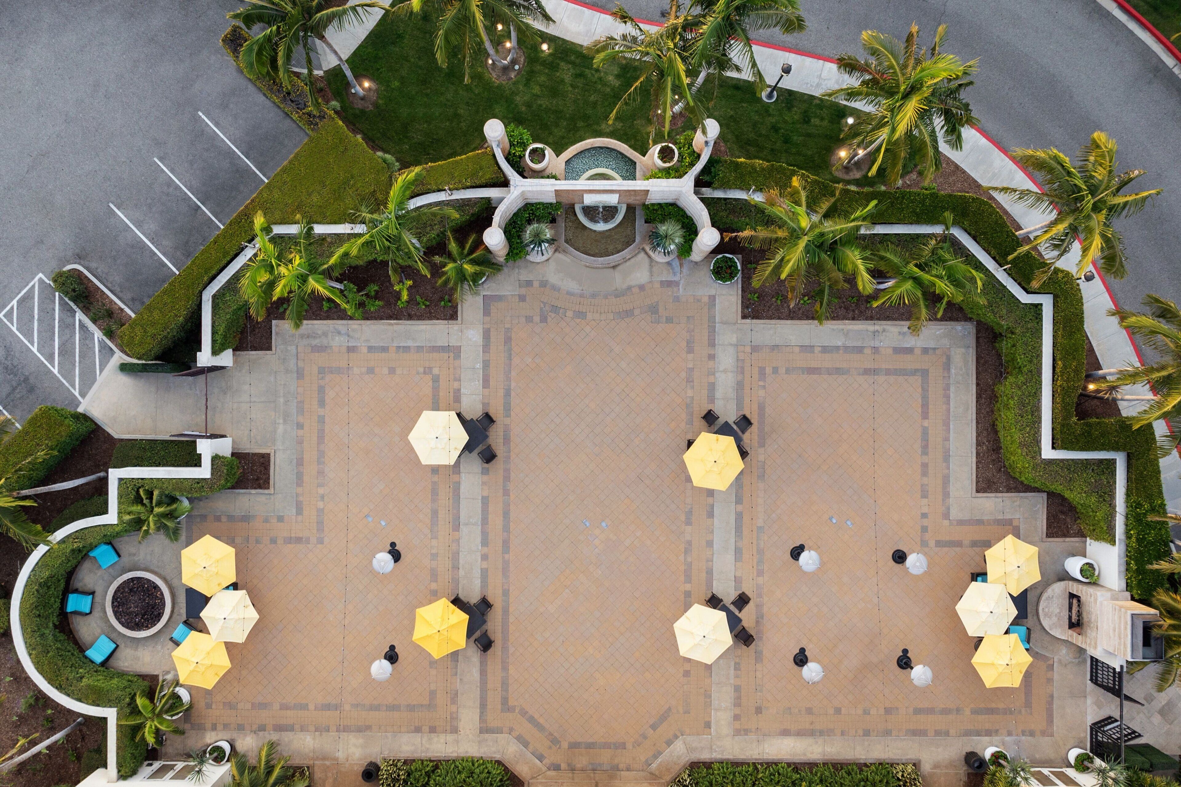 An aerial view of a courtyard with a central fountain, surrounded by seating areas with umbrellas, palm trees, and a parking lot.