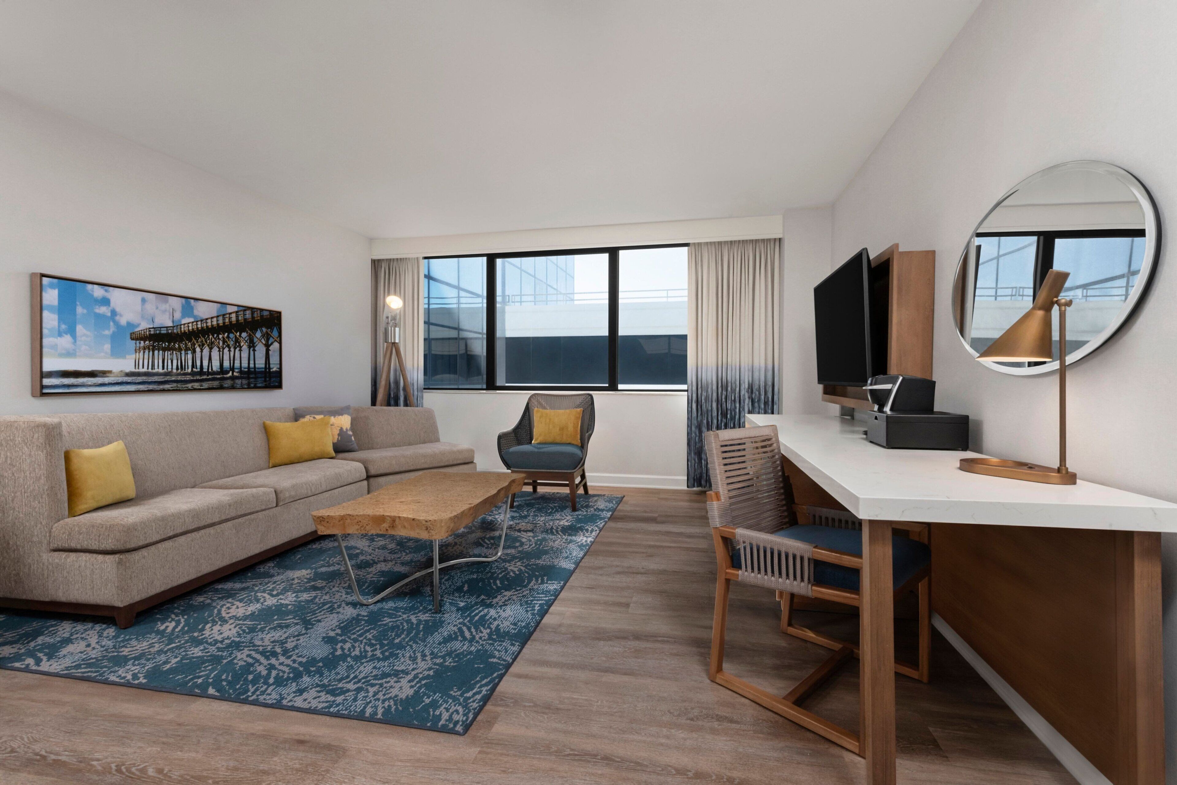 Een moderne hotelkamer met een bank, fauteuil, salontafel, bureau en grote ramen met uitzicht op de stad.