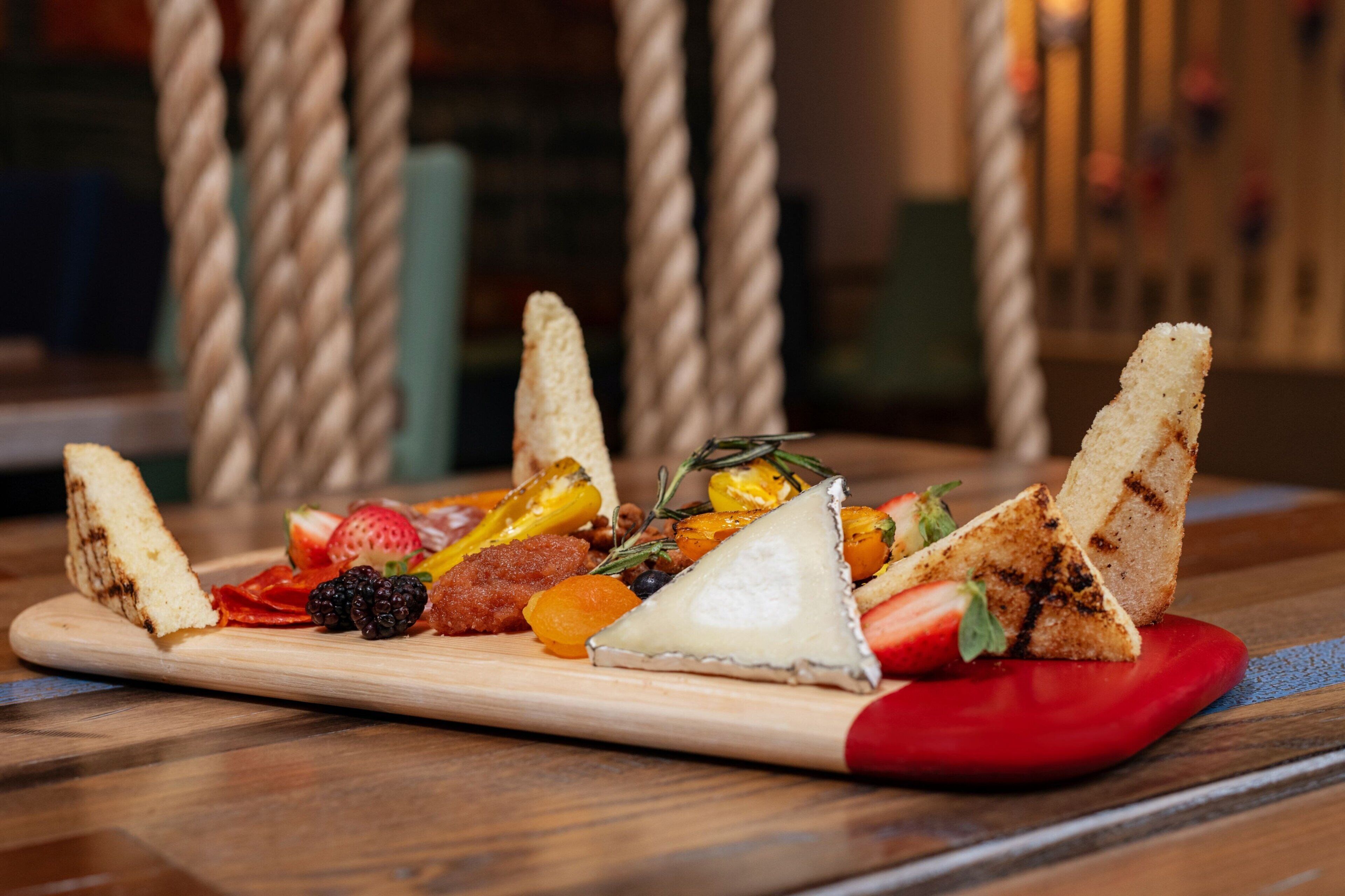 Una tabla de madera con varios quesos, frutas y pan sobre una mesa.