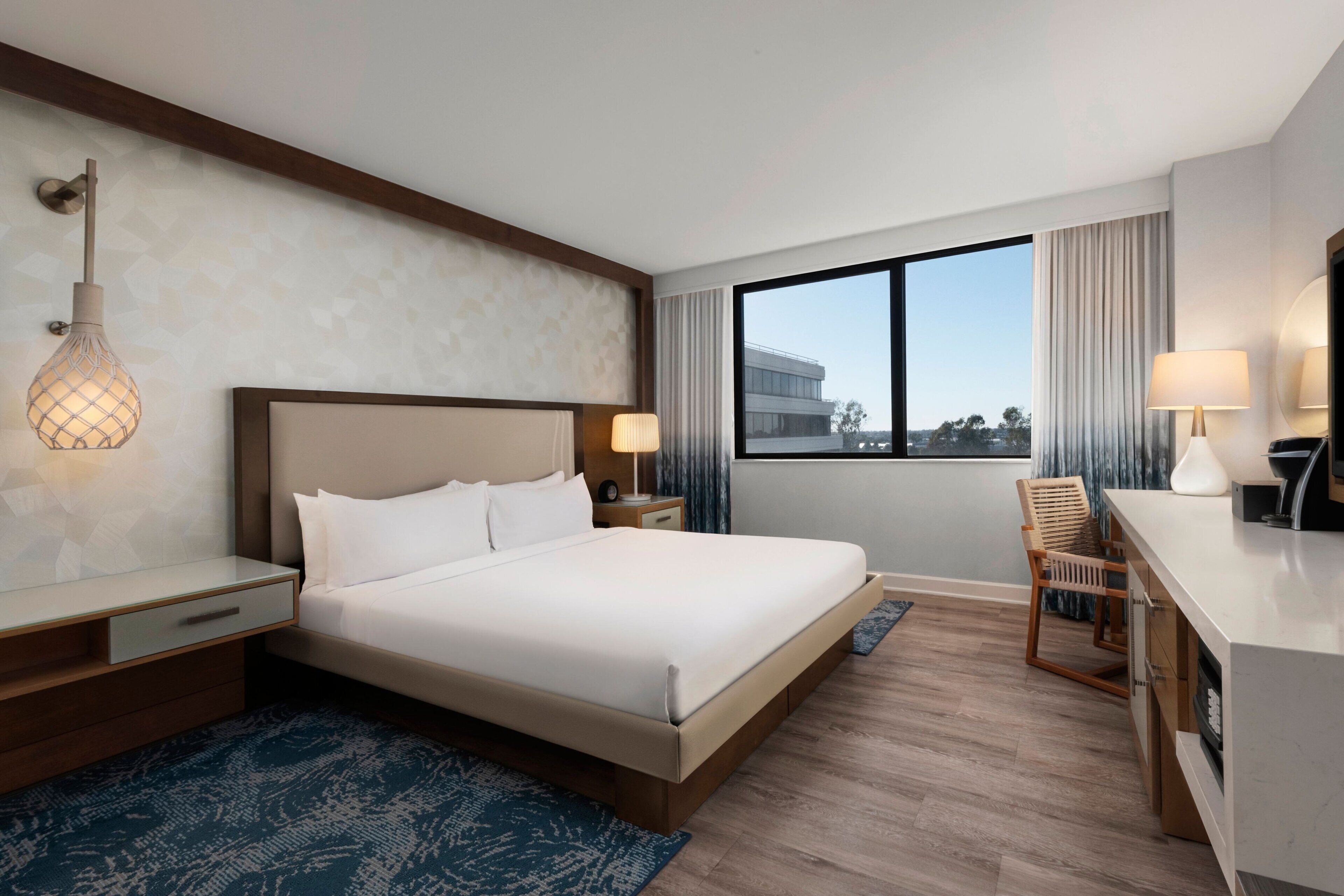 A hotel room with a large bed, bedside tables, a desk, and a chair. There is a window with curtains and a view of buildings outside.