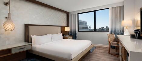 A hotel room with a large bed, bedside tables, a desk, and a chair. There is a window with curtains and a view of buildings outside.