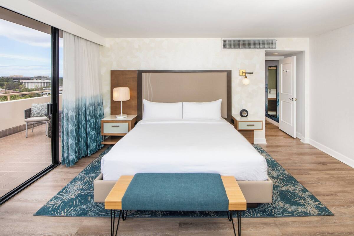Una habitación de hotel moderna con una cama grande, una mesita de noche con lámpara, un banco y vistas a la ciudad a través de amplios ventanales.