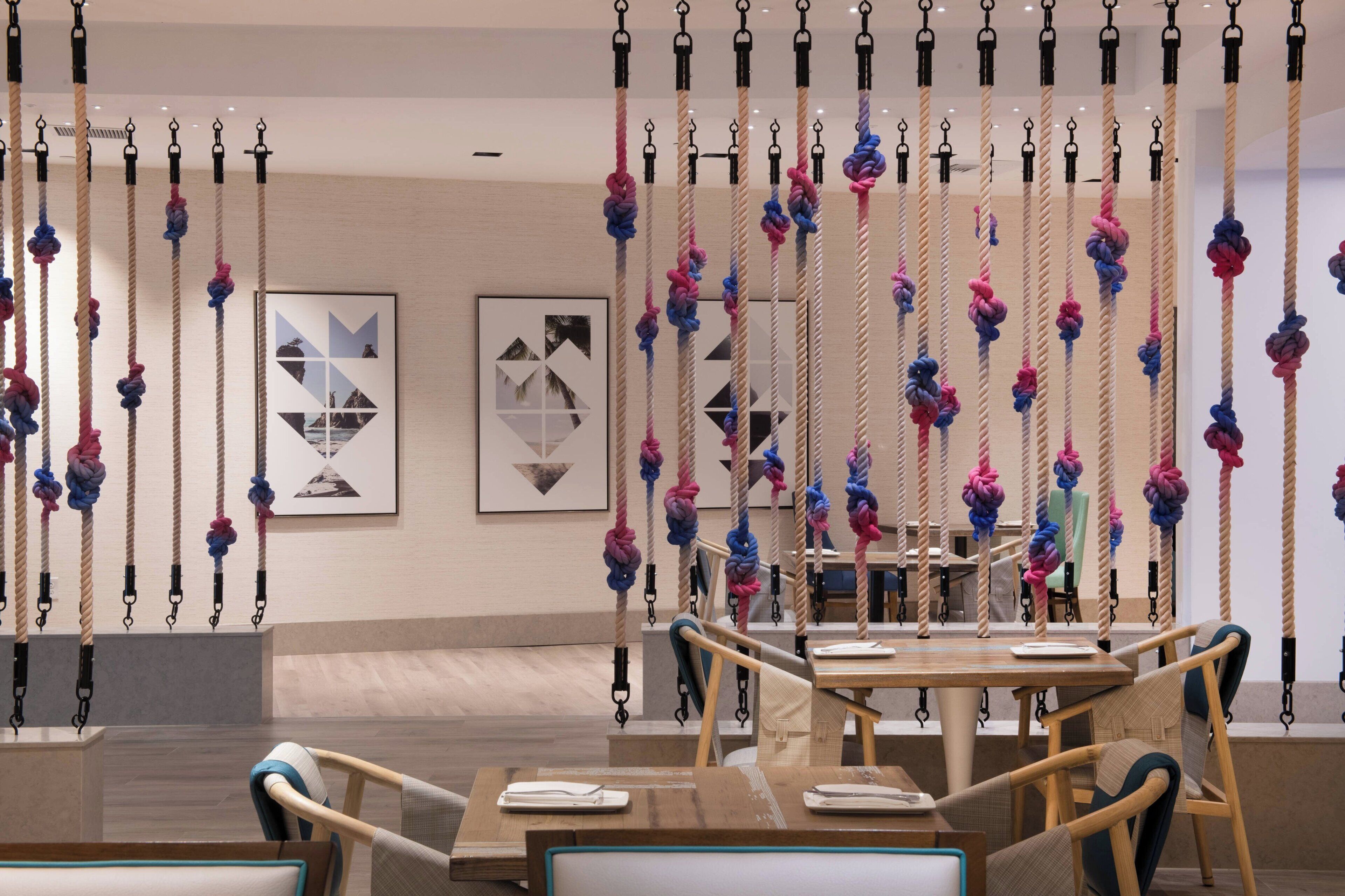 Een modern interieur met een eethoek, met houten tafels en stoelen, een muur met abstracte kunst en touwen die van het plafond hangen.