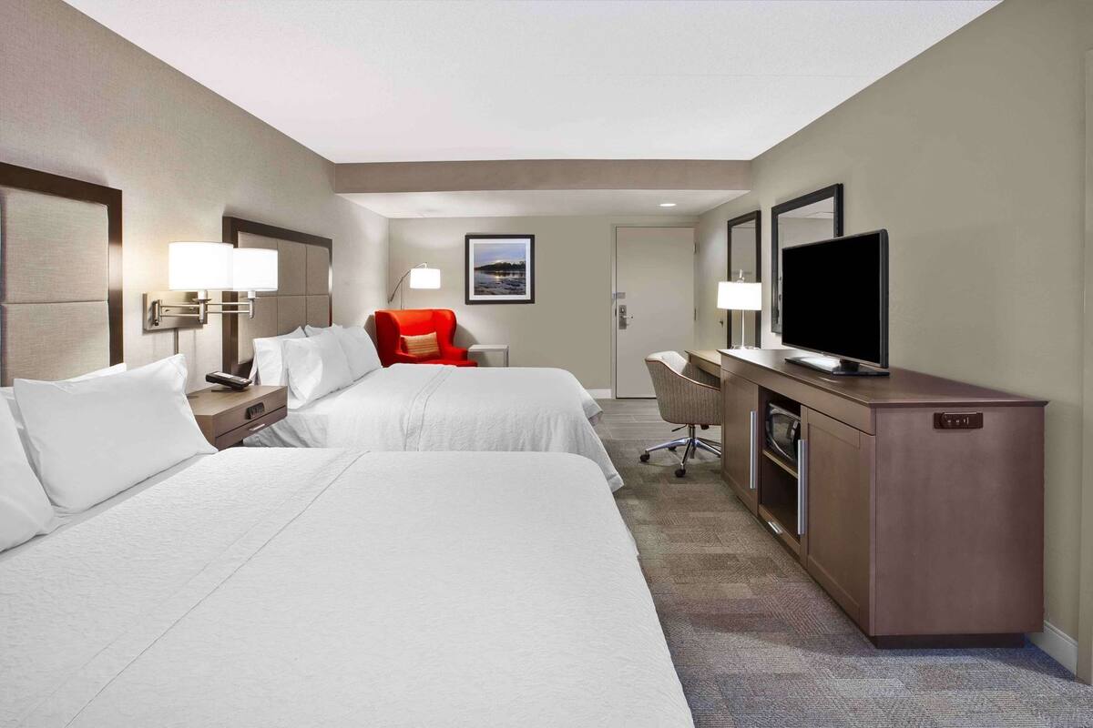 Quarto de hotel com duas camas, uma escrivaninha, uma TV e uma cadeira vermelha.
