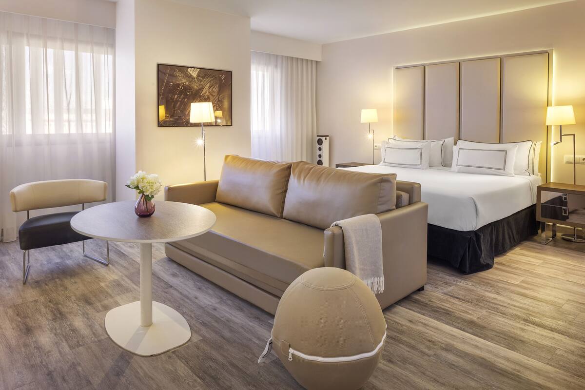A modern hotel room with a sofa, a round table, a bed with white linens, and a painting on the wall.