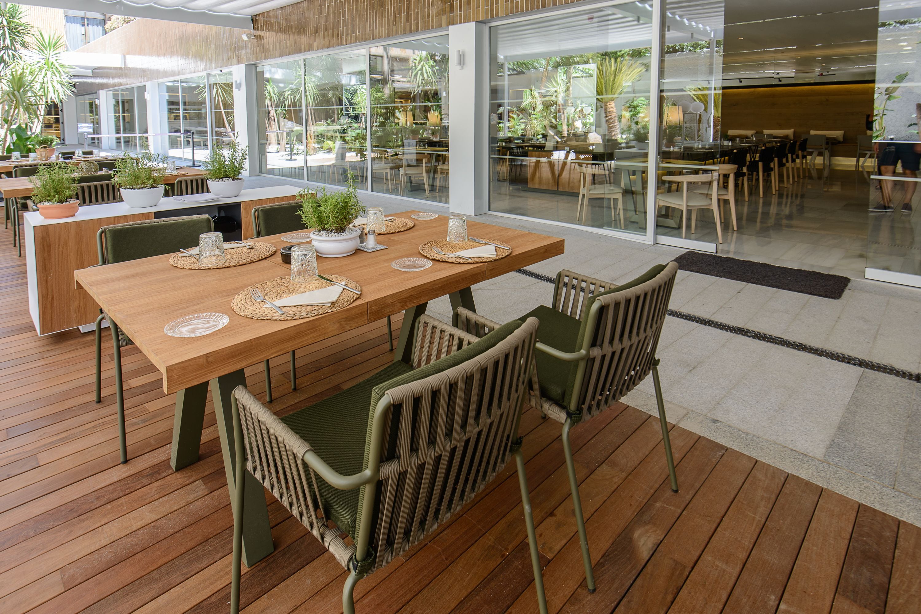 Un área de comedor al aire libre con mesas y sillas de madera, plantas en macetas y vistas del entorno verde.