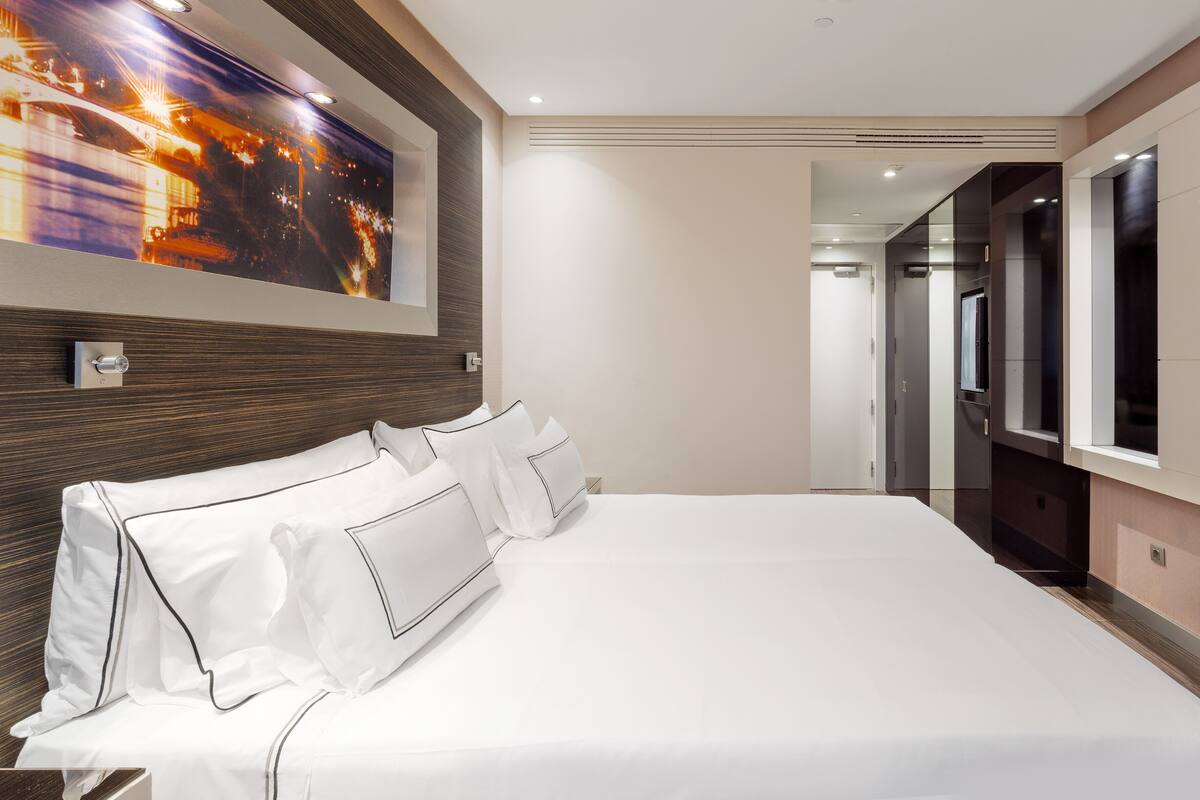 A modern hotel room with a large bed, a night view painting, and a hallway leading to a bathroom.