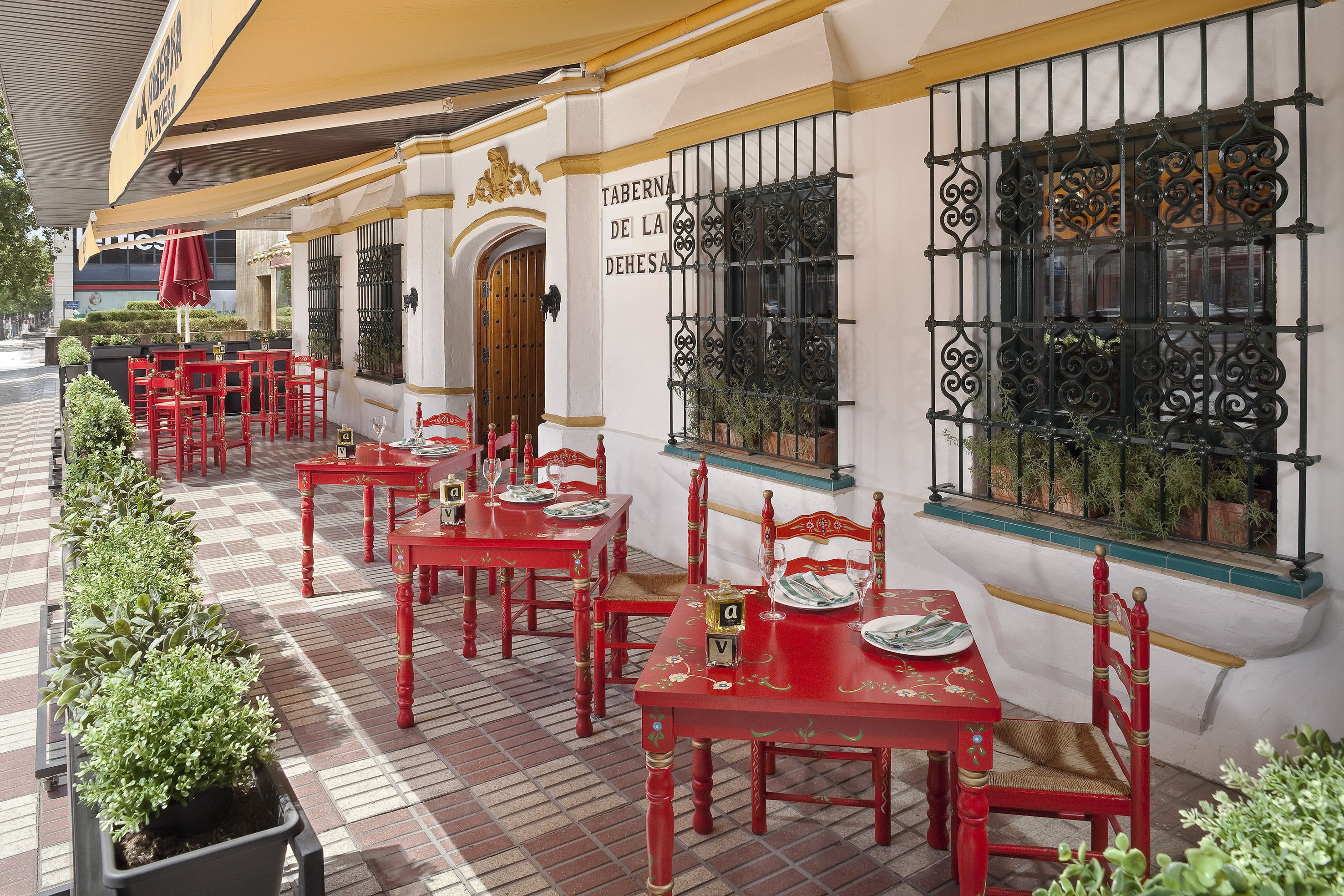 An outdoor dining area with red tables and chairs, a checkered floor, and a sign reading "TABERNAS DE LA DEHESA".