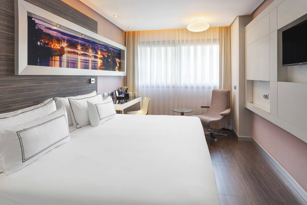 A hotel room with a large bed, a desk, a chair, a television, and a window with curtains.