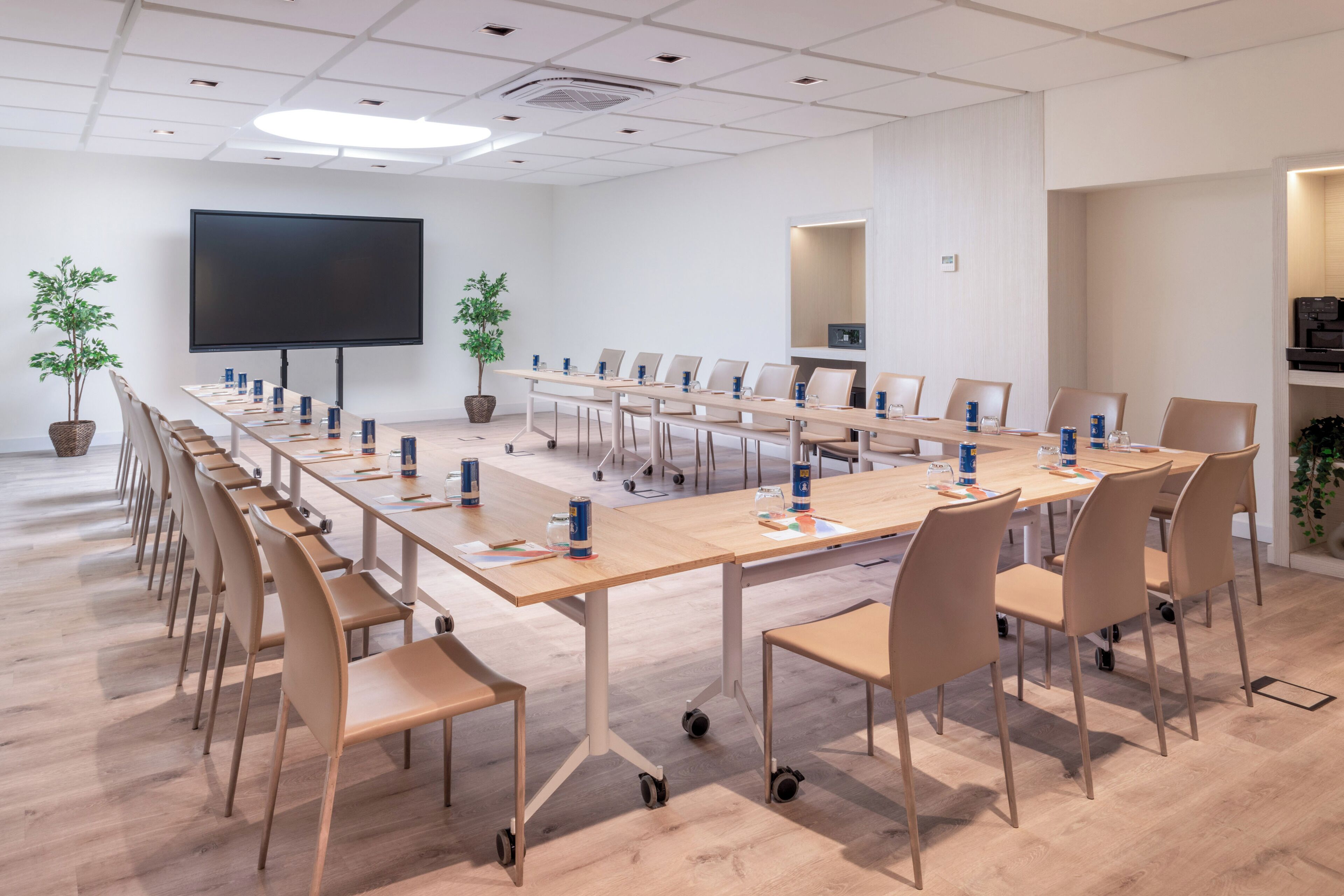 Un salón de conferencias con una larga mesa, sillas, una pantalla de proyector y plantas en macetas.