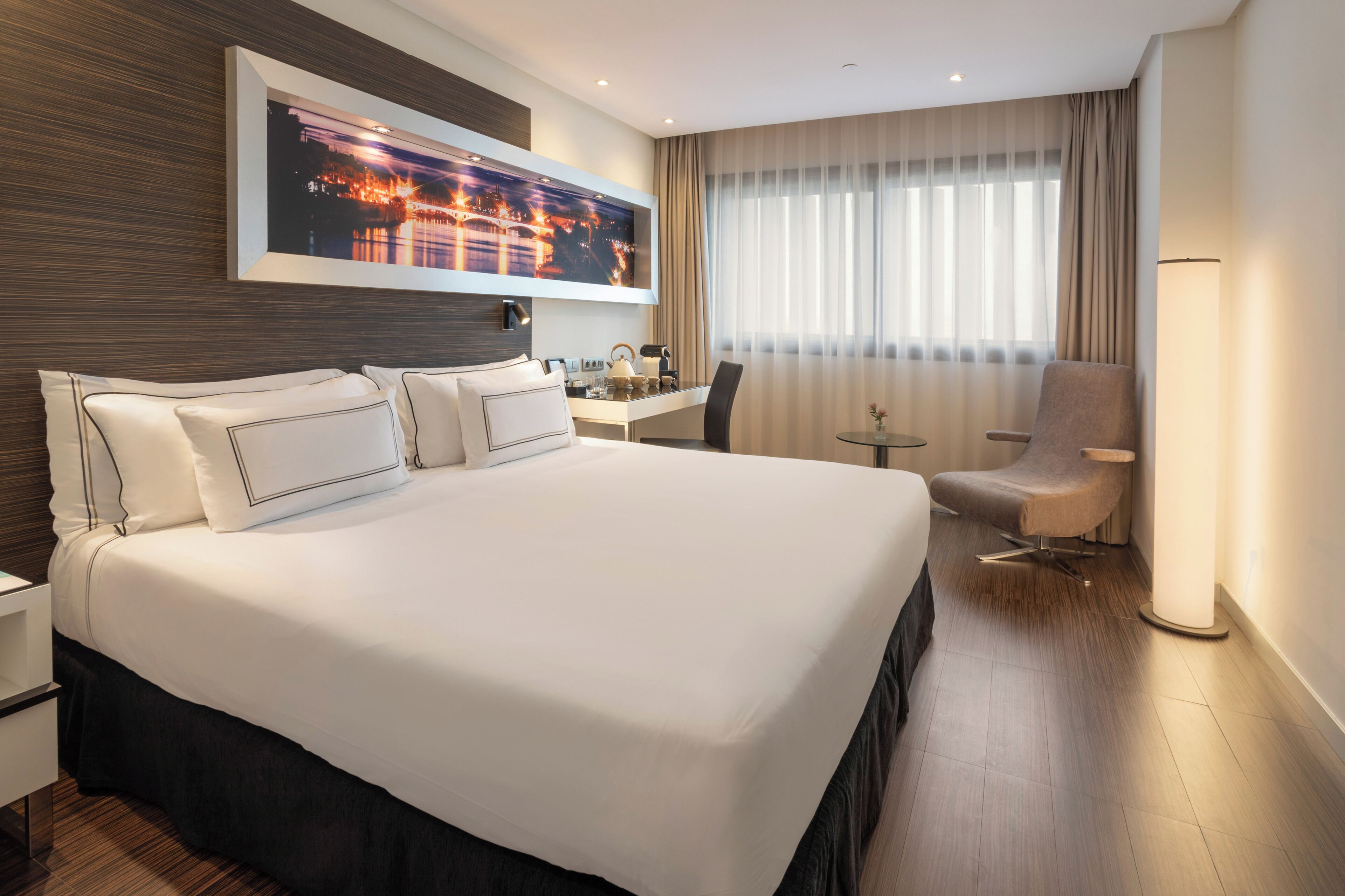 A hotel room with a large bed, a desk, a chair, and a large window with curtains.