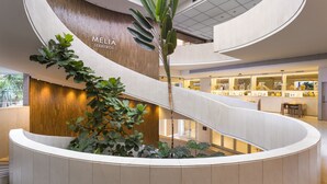 Un interior moderno con un mostrador de recepción curvo, una pared de madera con la señalización 'MELIA LEBRELOS' y una escalera que conduce a niveles superiores.