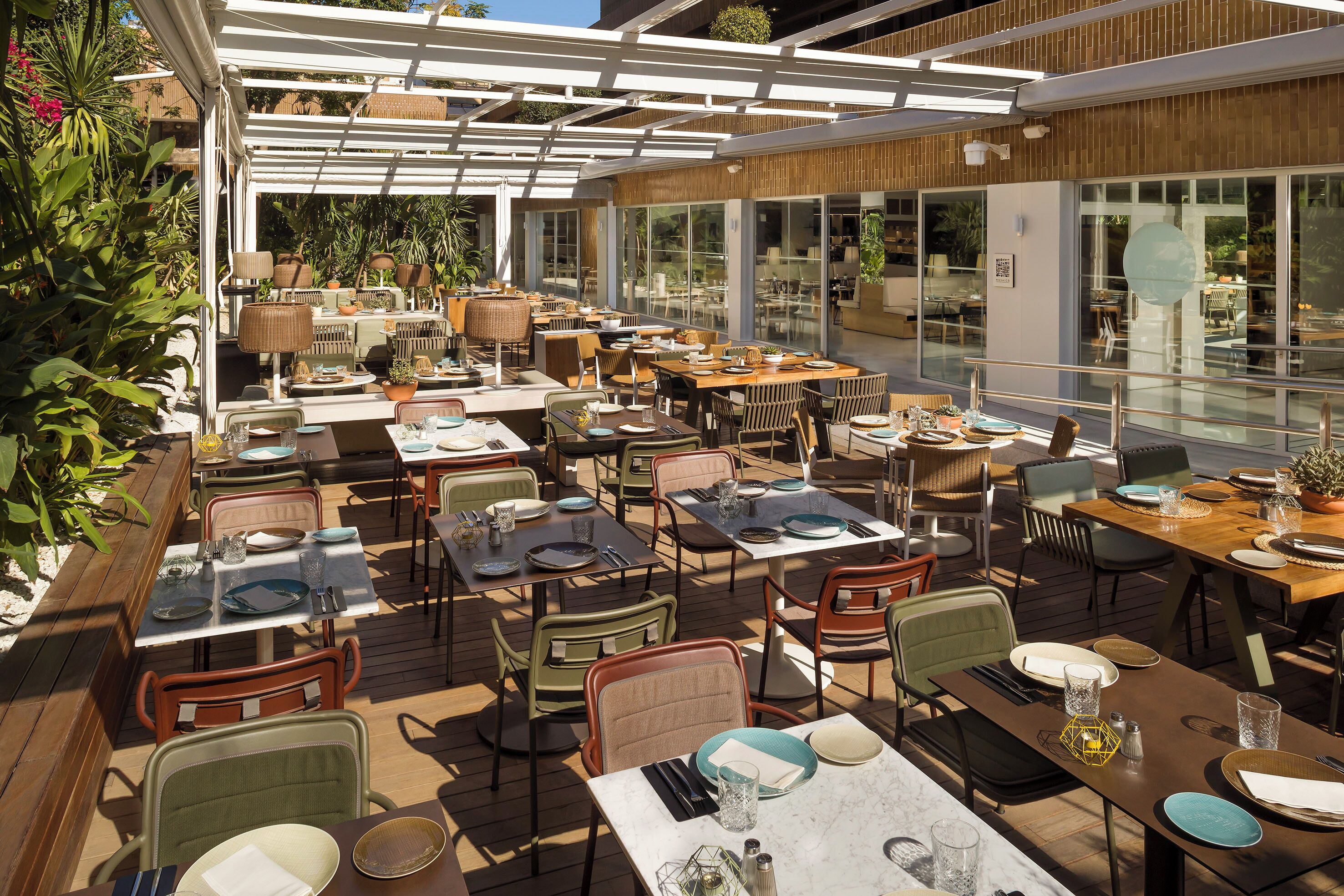 An outdoor dining area with multiple tables and chairs, covered by a pergola, surrounded by plants and a modern building in the background.